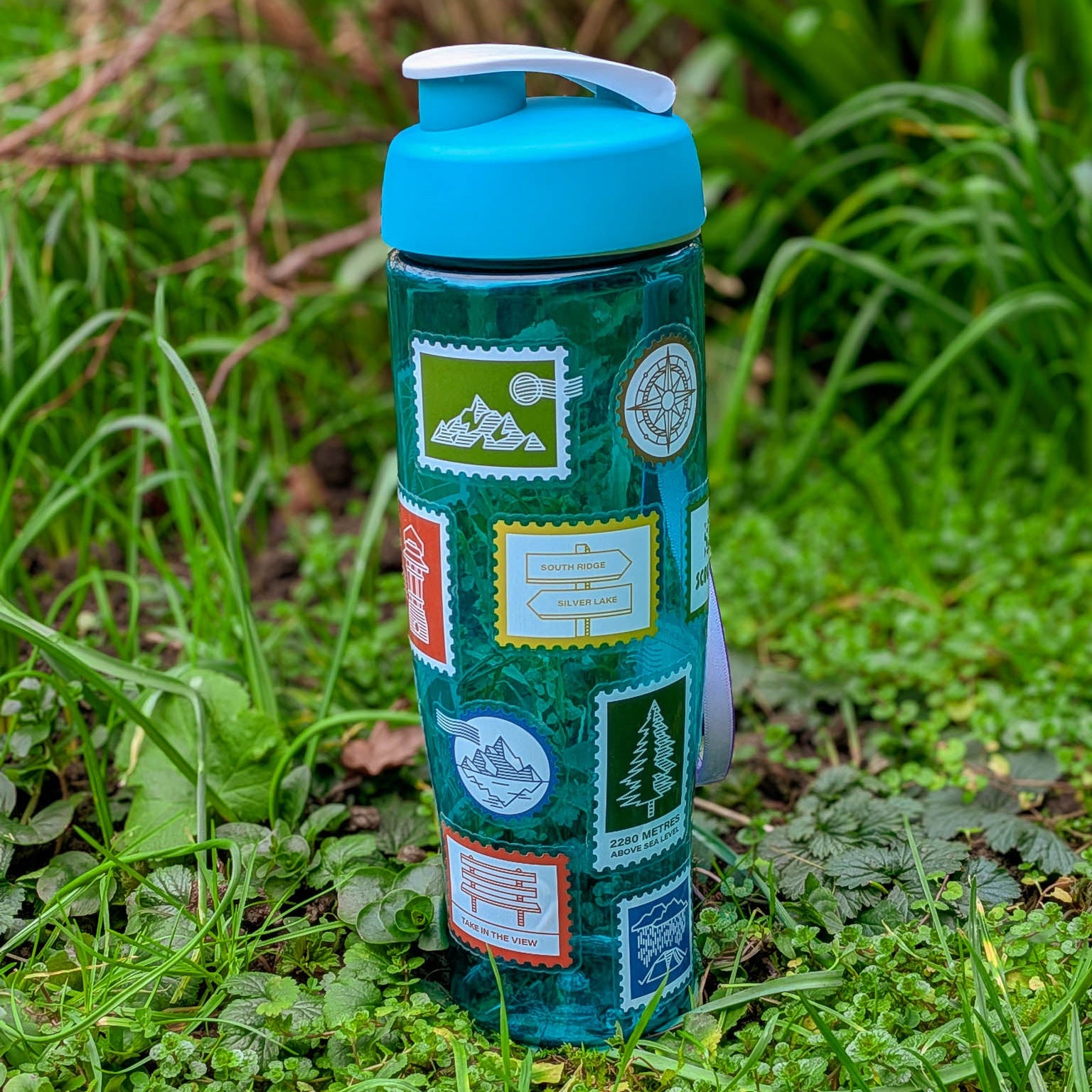 Blue water bottle with stamp-shaped stickers on a grassy background. Stickers are hiking, camping, scout and mountain themed.