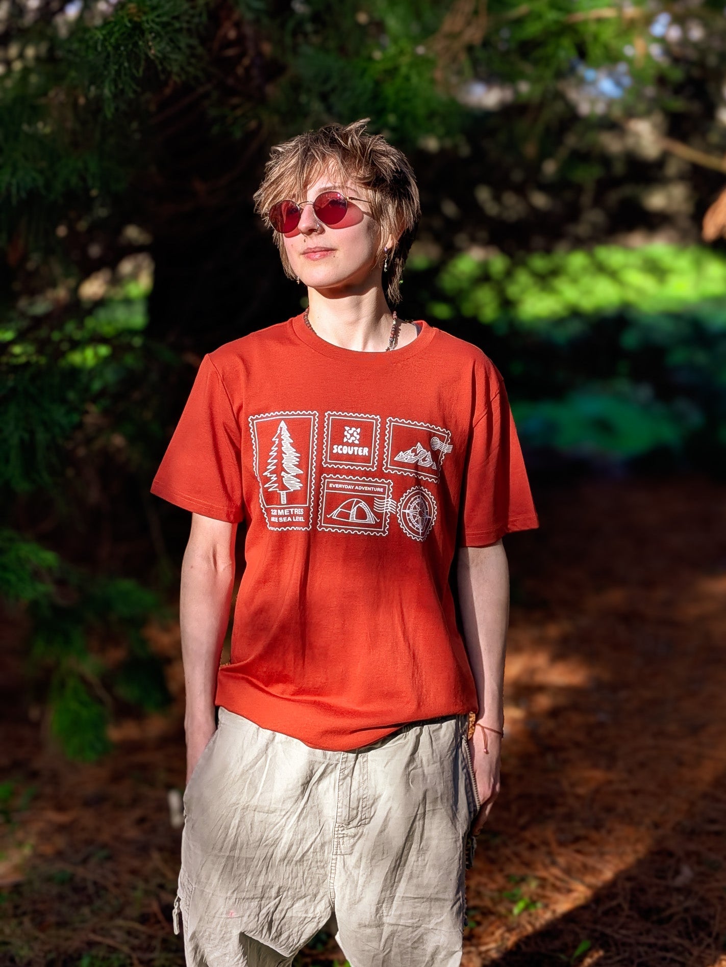 Person wearing a red t-shirt with graphics and light-colored pants standing in a forest.