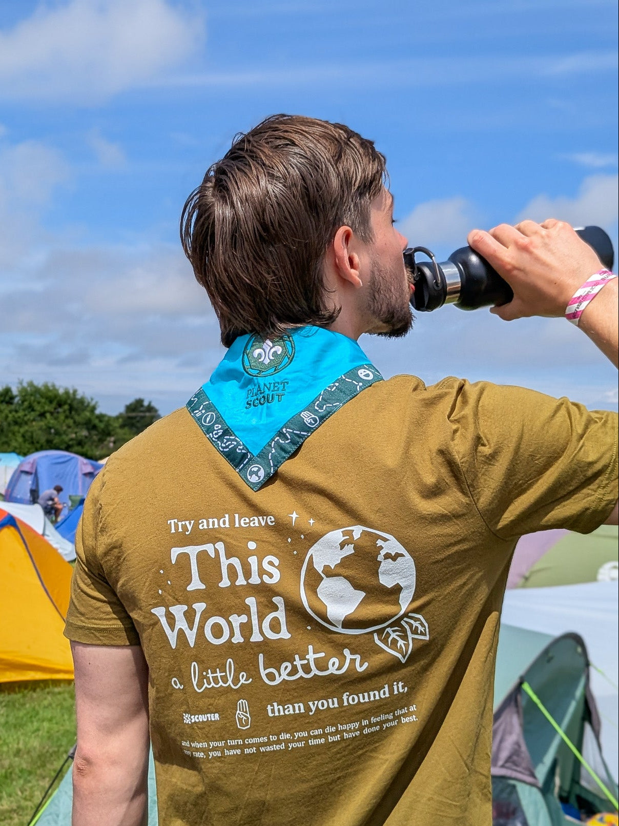 A man at an outdoor camping festival, seen from behind, wearing an olive green t-shirt with a large white graphic on the back that reads 'Try and leave This World a little better than you found it' with a globe illustration. He is wearing a blue scout scarf that read 'Planet Scout'. He is drinking from a black bottle, with colorful tents and a blue sky in the background.
