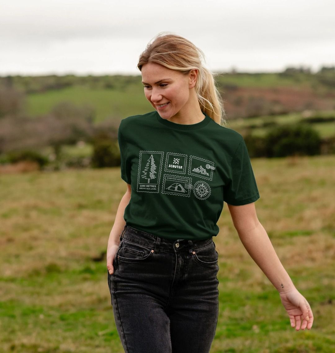 Woman walking through a field wearing evergreen coloured unisex t-shirt. Print design on tee shows a pine tree, mountains, compass, tent and Scouter logo displayed as stamps. Graphic is white line art.