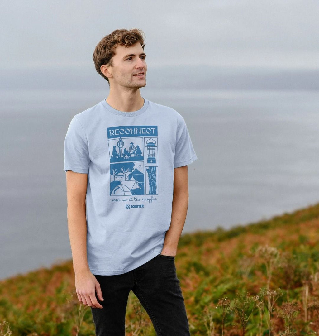 Man wearing light blue t-shirt, standing in a field. T-shirt features large blue print of Scouts around a campfire, a campsite with tents, a camp lantern and trees. Top text reads 'Reconnect'. Bottom text in cursive reads 'Meet me at the campfire'. Scouter logo sits at the bottom of the design.