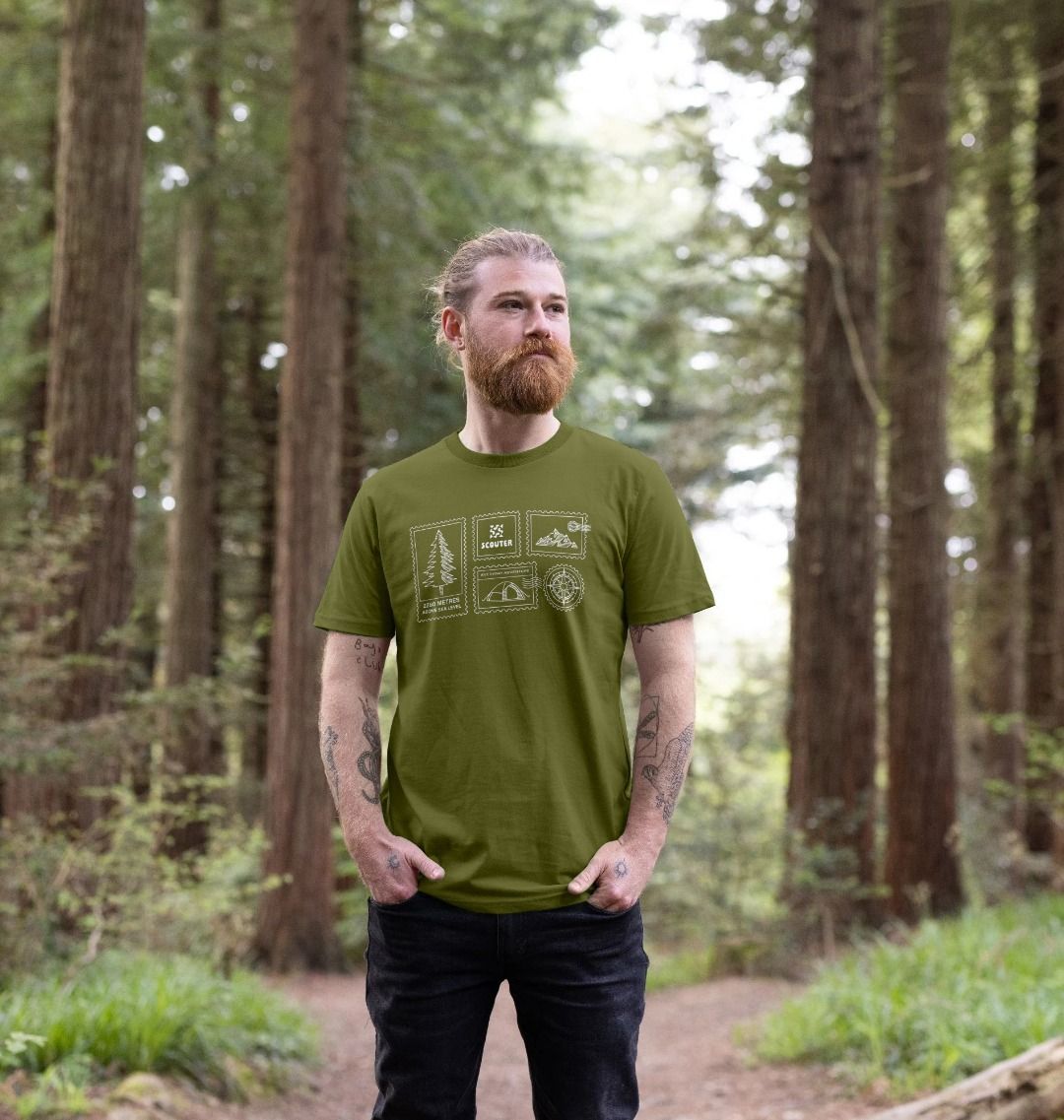 Man in a forest wearing moss green t-shirt coloured unisex t-shirt. Print design on tee shows a pine tree, mountains, compass, tent and Scouter logo displayed as stamps. Graphic is white line art.